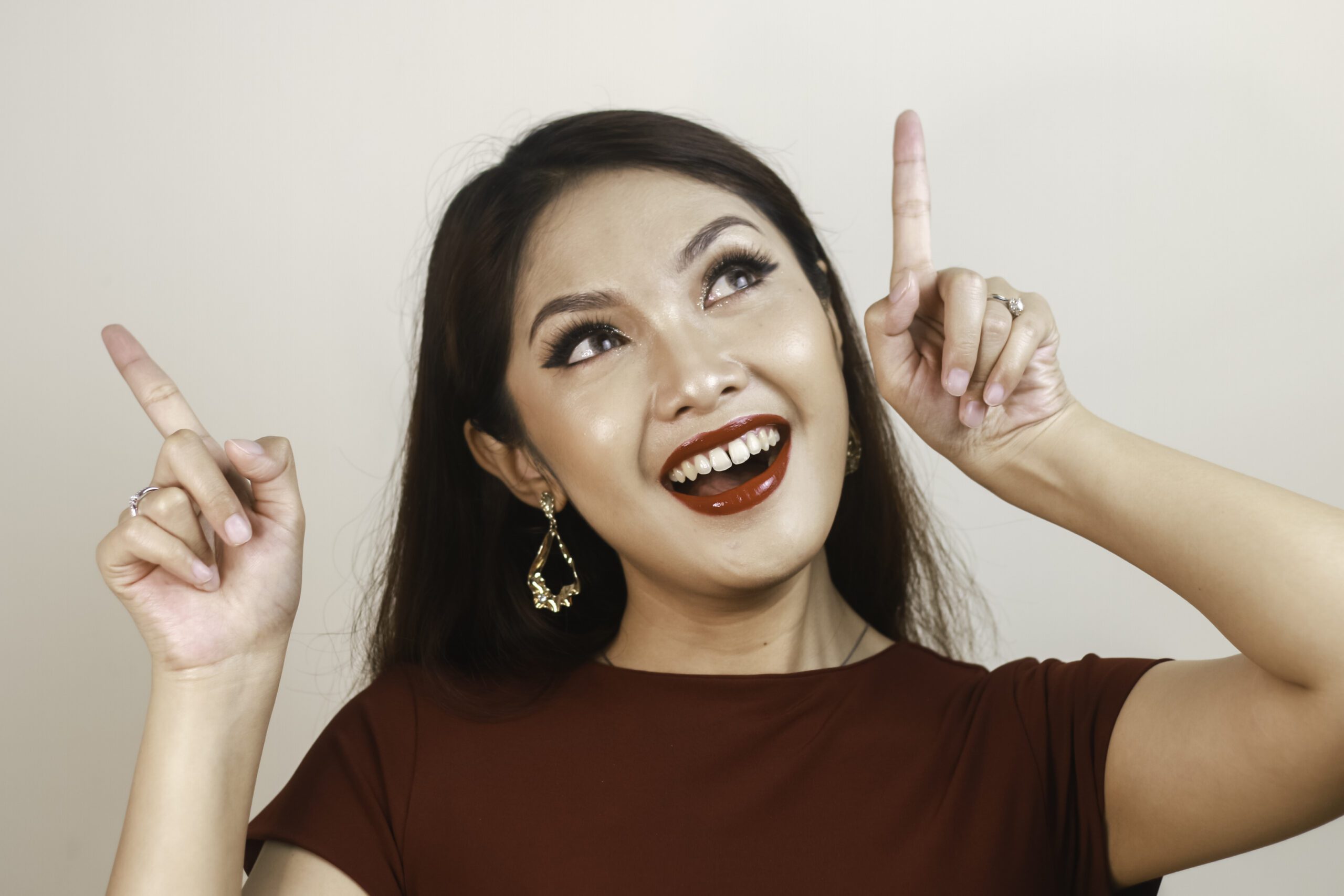 Shock face of a young Asian woman wearing a red shirt pointing upwards. Advertisement concept.